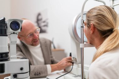 Optometrist aklinik göz muayenesi için keratografı ayarlıyor. Masada oturan kadın ve erkek birbirleriyle konuşuyorlar..