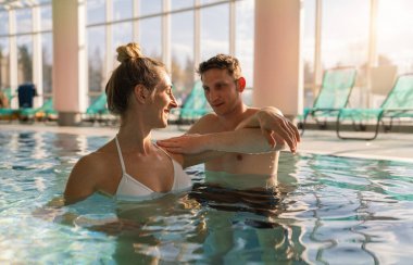 Beyaz mayo giymiş bir kadın spa merkezindeki bir havuzda omuz egzersizi yaparken bir erkek eğitmene gülümsüyor. aqua aerobik konsept resmi