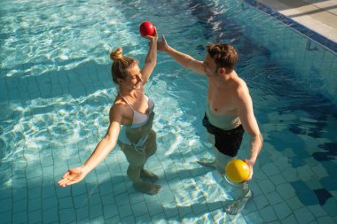 Havuzdaki kadın ve erkek güneşli bir rehabilitasyon seansında suyun üzerinde renkli egzersiz toplarını kaldırıyor.