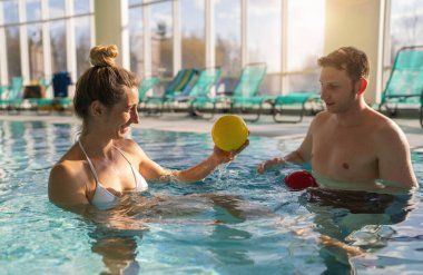 Kadın müşteri ve erkek antrenör gülümsüyor ve kapalı bir havuzda renkli egzersiz topları kullanıyorlar.