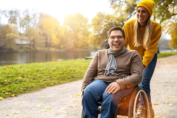 Happy Woman and her smiling friend in a wheelchair having stroll through the Riverside at Park in autumn. Supportive Moments concept image