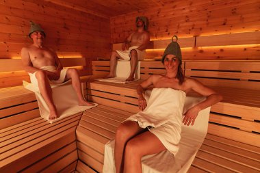 Saunada üç kişi keçeli şapka takıyor, iki erkek oturuyor ve bir kadın yaslanıyor.