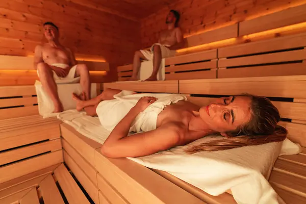 Reclining woman and seated men in a wooden Finnish sauna, warm ambient light