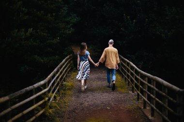  A romantic photo of a young couple in a beautiful autumn park, walking hand in hand down a pathway surrounded by colorful foliage. The couple is lost in their own world as they hold hands and stroll down the path, enjoying each other's company and t