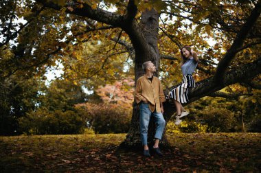 Güzel bir sonbahar parkında genç bir çiftin romantik bir fotoğrafı. Ağacın yanında duran ve ağaç dalında oturan sevgilisine bakan bir adam. Çift bir arkaplan ile çevrili