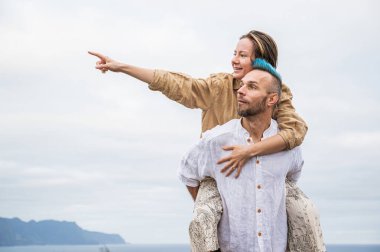 A young couple walks on a road surrounded by nature, with the man carrying the woman on his back. They appear to be taking in the beauty of their surroundings and enjoying the moment together. Both