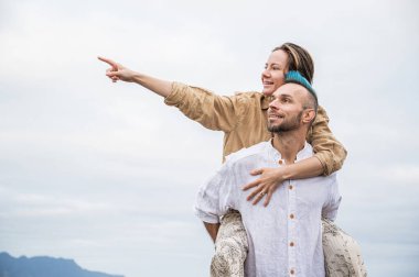 A young couple walks on a road surrounded by nature, with the man carrying the woman on his back. They appear to be taking in the beauty of their surroundings and enjoying the moment together. Both