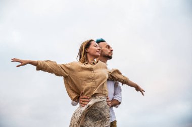 A young couple stands with their arms open and eyes gazing into the distance on a rocky cliff by the ocean. The man has his hands on the womans waist, and she has her arms outstretched. The couple