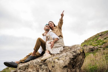 A young couple sits on a rock surrounded by hills and rocks in the background. They appear to be taking in the natural beauty of their surroundings and enjoying each others company. The couple sits
