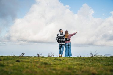 A romantic and intimate photo of a couple in nature, with a clear blue sky as the background. The male partner holds the female partner, embracing her tenderly as she points with her hand in the far