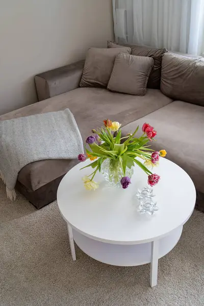 Image of beige soft sofa with white round wooden table and colourful flowers in the glass vase
