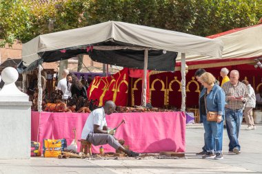 ALCALA de HENARES, MADRID, SPAIN - 11 Ekim 2022: Turistler atölyesinde çalışan ve ahşap oymalar satan Afrika kökenli genç bir heykeltıraşı izliyorlar. Şehrin Cervantino pazarının kutlaması sırasında çekildi.