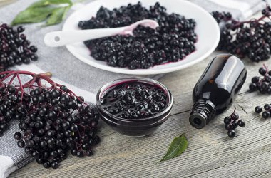 Homemade black elderberry syrup in ceramic jar and bunches of black elderberry with green leaves on wooden desk.