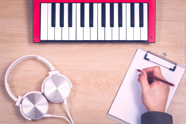 曲作りのコンセプト 木製の机の上に赤い音楽キーボードと白いヘッドフォンで紙に曲を書くクローズアップソングライターの手 — ストック写真