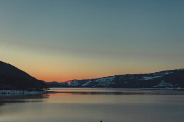 Sakin bir göle yansıyan karlı dağların üzerinde sakin bir günbatımı. Gökyüzü ılık renklerle parlıyor, kış manzaralarını güzelleştiriyor..