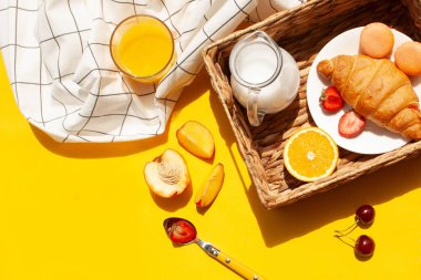 Hasır sepet kruvasanlarla sabah kahvaltısı, sarı arka planda süt ve meyveler
