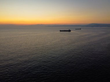 Ufukta iki gemi olan bir gün batımının fotorealistik görüntüsü. Gökyüzü turuncu ve sarı, güneş ufukta batıyor. Okyanus koyu mavi ve sakindir..