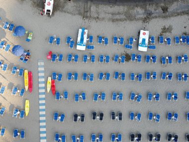 Temiz düzenlenmiş mavi şezlonglar, şemsiyeler, renkli kanolar ve cankurtaran istasyonları olan bir sahil manzarası.
