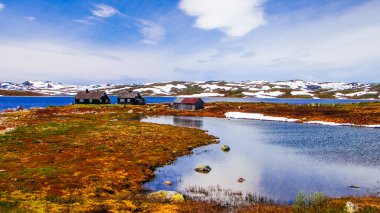 Eski ahşap evler, Norveç kırsalında mavi göl ve karla çevrili arazide yapayalnız duruyor.