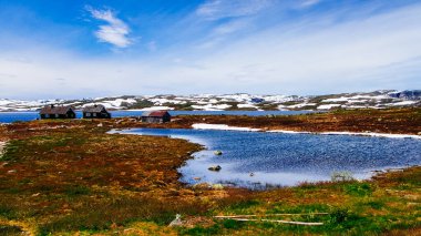 Eski ahşap evler, Norveç kırsalında mavi göl ve karla çevrili arazide yapayalnız duruyor.