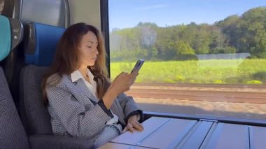 Trende oturan güzel bir kadın, dinleniyor ve akıllı telefon kullanıyor. Yaratıcı kız sosyal medyayı kontrol ediyor, arkadaşlarıyla sohbet ediyor, internette geziniyor. Yüksek kalite 4k görüntü