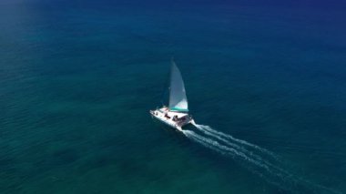 Havadan çekilen görüntüler, açık mavi denizde seyreden lüks yelkenli görüntüsü. Kamera mavi okyanus suyunda katamaranı takip ediyor..