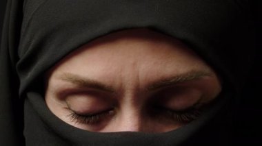 Close-up portrait of a Muslim woman with beautiful eyes in a hijab on a dark background. A girl in a black headscarf is looking at the camera. Arab women life concept. High quality 4k footage