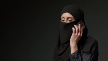 Woman in black muslim headscarf talking on mobile phone. Young arab muslim girl in headscarf hijab using smart phone, close up portrait of smiling middle eastern girl holding mobile smartphone.