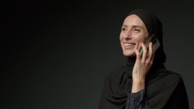 Woman in black muslim headscarf talking on mobile phone. Young arab muslim girl in headscarf hijab using smart phone, close up portrait of smiling middle eastern girl holding mobile smartphone.