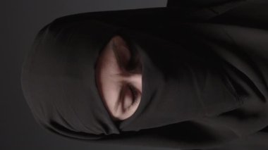 Vertical video of close-up portrait of a Muslim woman with beautiful eyes in a hijab on a dark background. A girl in a black headscarf is looking at the camera. Arab women life concept.