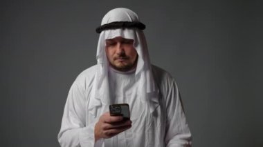 Authentic muslim businessman in traditional white kandura with beard using smartphone. Successful Saudi, Emirati, arab businessman concept. High quality 4k footage