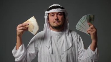 Successful Arabic sheikh in traditional clothes holds money in his hands on a gray background. Rich Arab man concept. High quality 4k footage
