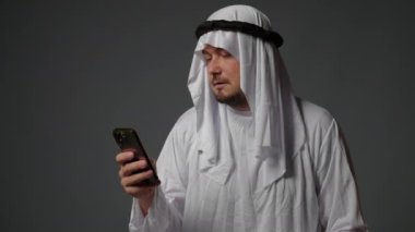 Authentic muslim businessman in traditional white kandura with beard using smartphone. Successful Saudi, Emirati, arab businessman concept. High quality 4k footage