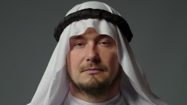 The sheikh wearing a white kandura and a gutra on his head close up looks at camera gray background. High quality 4k footage