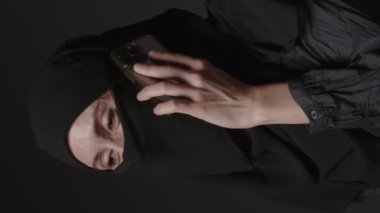 Vertical video of woman in black muslim headscarf talking on mobile phone. Arab muslim girl in headscarf hijab using smart phone, close up portrait of smiling middle eastern girl holding smartphone.