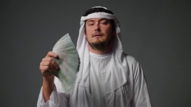 Successful Arabic sheikh in traditional clothes holds money in his hands on a gray background. Rich Arab man concept. High quality 4k footage