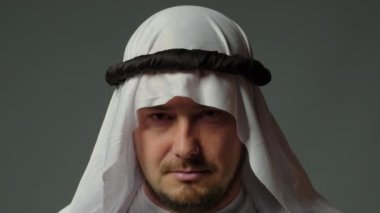 The sheikh wearing a white kandura and a gutra on his head close up looks at camera gray background. High quality 4k footage