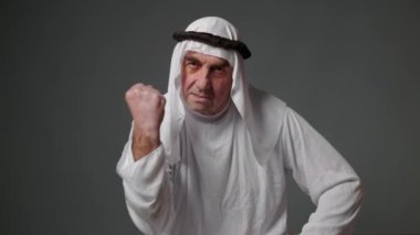 An adult Muslim man in white traditional clothes looks at the camera and expresses emotions. Elderly male sheikh on a gray background. High quality 4k footage