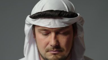 The sheikh wearing a white kandura and a gutra on his head close up looks at camera grey background. High quality 4k footage