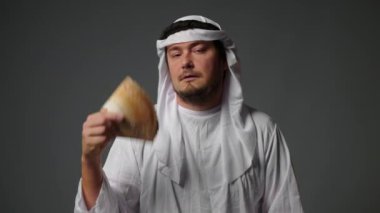 Successful Arabic sheikh in traditional clothes holds money in his hands on a gray background. Rich Arab man concept. High quality 4k footage