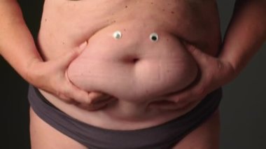 Göbek yağlı kadın. Yaşlı kadınlarda obezite. Şişman kadın göbeği, selülitleri kontrol etmek için deriyi tutan kadın. Şişman bir karında sarkık deri, gri arka planda plastik cerrahi konsepti. Yüksek kalite 4k görüntü