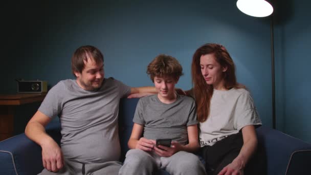一家人坐在家里一个漂亮的室内蓝色沙发上 一边看着电话 一边带着愉快的心情笑着 青少年的对话和教育 良好的家庭关系 — 图库视频影像