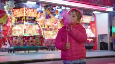 Küçük bir kız, renkli ışıkların arka planında neşeli bir çocuk etkinliğinde pembe pamuk şeker yiyor. Çocuklar için tatil eğlencesi. Yüksek kalite 4k görüntü