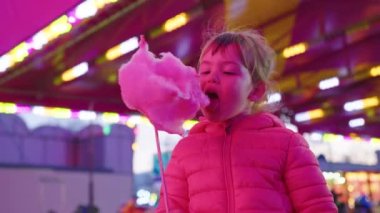 Küçük bir kız, renkli ışıkların arka planında neşeli bir çocuk etkinliğinde pembe pamuk şeker yiyor. Çocuklar için tatil eğlencesi. Yüksek kalite 4k görüntü