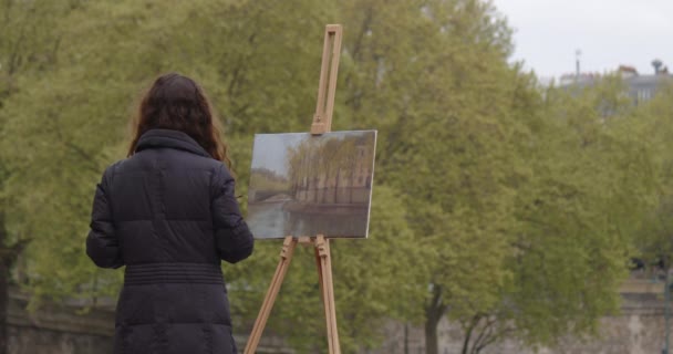 セーヌ川の近くで絵を描く女性 パリのイーゼルの近くに立っている間 クリエイティブな女性アーティストがブラシで絵を描きます 女性アーティスト 販売のための芸術 インスピレーション — ストック動画