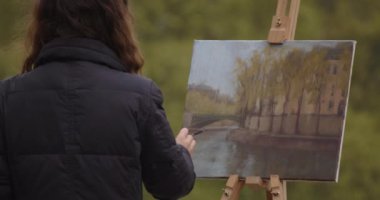 Kadın sokak ressamının Paris şehir manzarası ile eskiz çalışması var. Deneyimli Avrupalı kadın sanatçı nehir kenarında yeni bir resim çiziyor. Ressam ve onun yaşam tarzı hobisi..