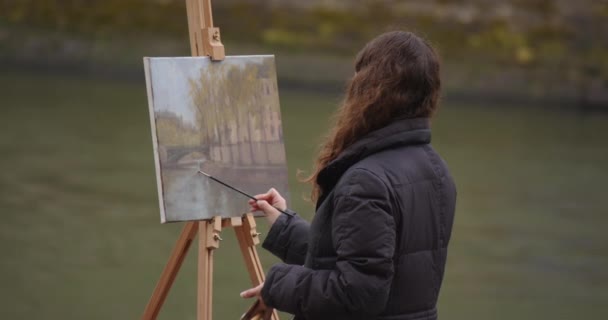 セーヌ川の近くで絵を描く女性 パリのイーゼルの近くに立っている間 クリエイティブな女性アーティストがブラシで絵を描きます 女性アーティスト 販売のための芸術 インスピレーション — ストック動画