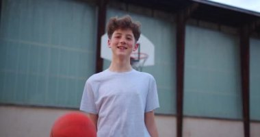 Şık bir saç stili olan genç bir adam kameraya bakıyor ve bir basketbol topuyla oynuyor. Okuldan sonra açık havada bir gencin portresi. Modern çocukların yaşam tarzı..