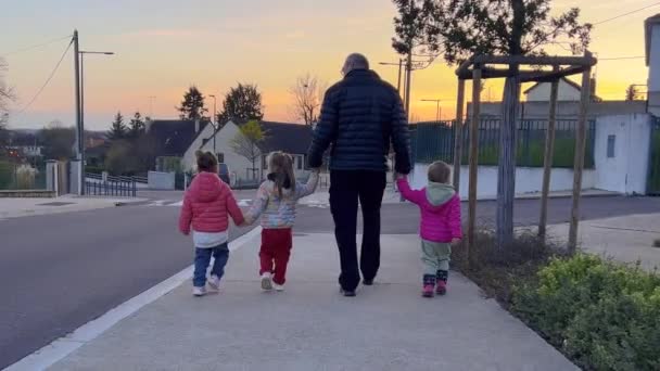3人の娘と街の通りを歩いている高齢者の父親のリアビュー 子供たちが手を握っている 家族は日没時に街中を歩く 小さな女の子はパパと外に出て — ストック動画