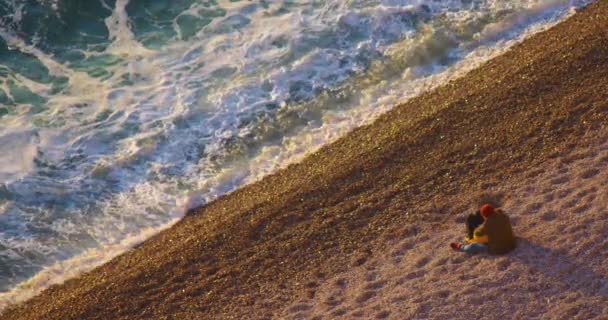 一对年事已高的夫妇坐在大海里欣赏日落时的海浪 恋爱中的快乐夫妻 日落时坐在沙滩上 旅行度假生活方式 法国的石滩 — 图库视频影像